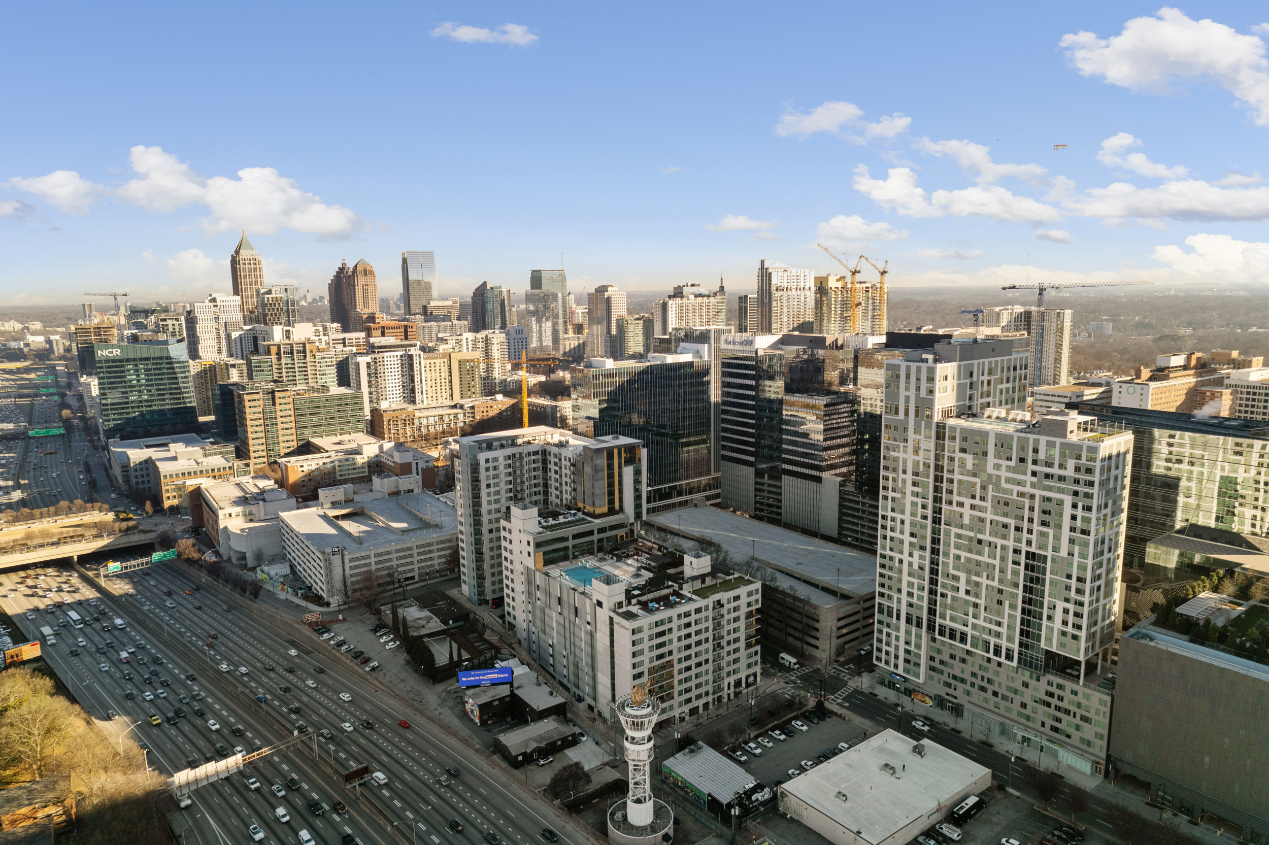 Aerial view of Midtown Atlanta showcasing modern high-rise buildings, busy I-75/I-85 Connector freeway, construction cranes, and the city's central business district under a clear blue sky. Captured for Jimmy Carey Commercial Real Estate, Atlanta Restaurant Business Broker.