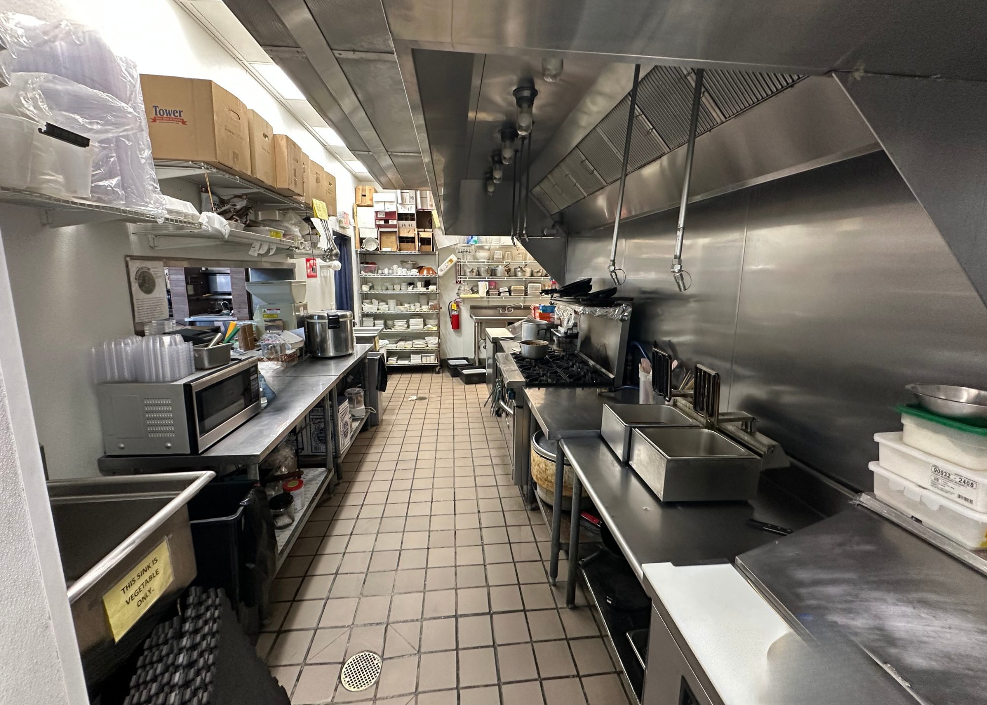 Interior of a fully equipped commercial kitchen inside a sushi restaurant for sale. Features stainless steel prep stations, wok range, fryer, industrial hood system, microwave, and organized shelving. Listed by Jimmy Carey Commercial Real Estate, Atlanta Restaurant Business Broker.