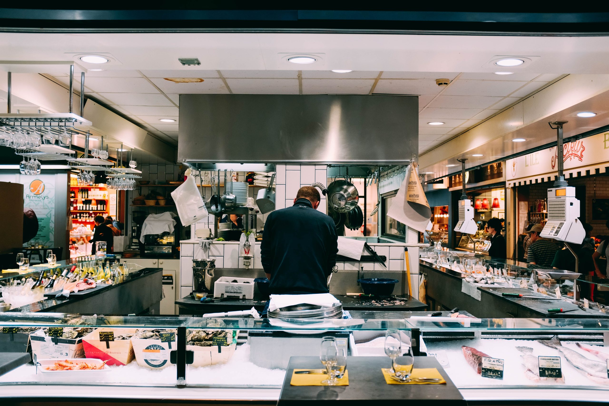Interior of a seafood market or open-concept restaurant kitchen with a chef preparing food behind a counter displaying fresh fish, oysters, and shellfish on ice. Hanging cookware, wine bottles, and glassware are visible, with customers and vendor stalls in the background. Captured for Jimmy Carey Commercial Real Estate, Atlanta Restaurant Business Broker.
